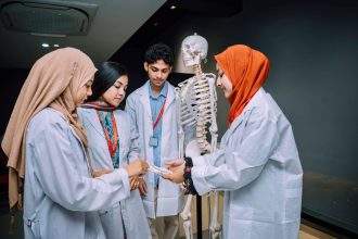 Grupo de estudantes de medicina estuda anatomia ao redor de um esqueleto em uma sala de aula.