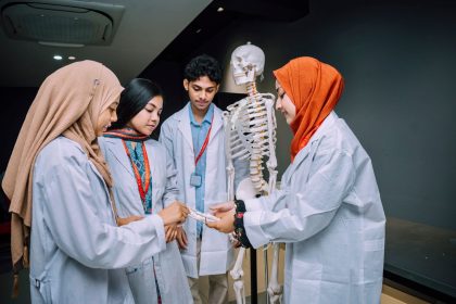 Grupo de estudantes de medicina estuda anatomia ao redor de um esqueleto em uma sala de aula.