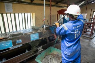 Trabalhador de macacão azul opera uma mesa vibratória para processamento de minério em uma instalação industrial.