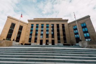 Fachada do prédio do Federal Reserve nos Estados Unidos, vista de baixo, com bandeiras americanas e céu nublado